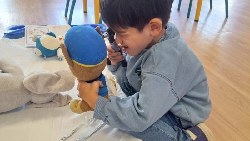 Un enfant ausculte sa peluche avec un otoscope, a cote du carnet de sante du nounours.